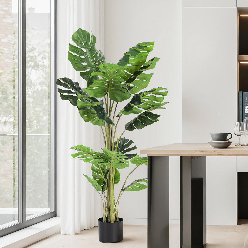 Artificial monstera plant in a modern interior setting with a table and window.